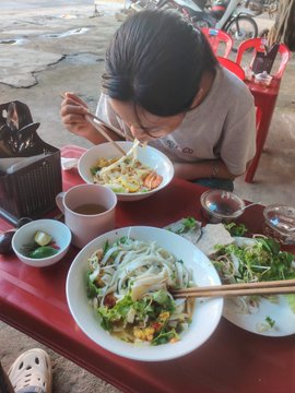 The image shows a person sitting at a table with two bowls of food - one containing noodles and another filled with salad. The individual appears to be eating from both bowls, enjoying their meal. There are also chopsticks present on the table for use during the meal.