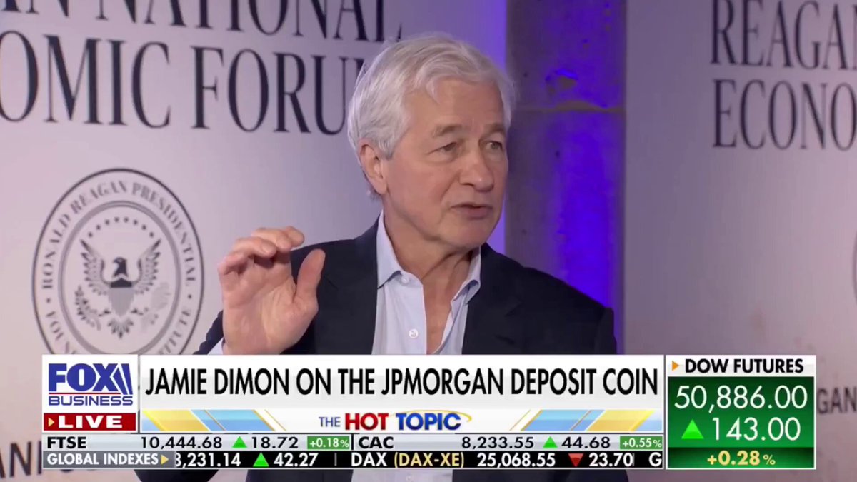 The image shows a man giving an interview, with his hand raised as if he's making a gesture or pointing at something. The background features a large screen displaying the news, which includes a headline about Jamie Dimon discussing the importance of the Clarity Act for banks and Bitcoin. The text on the screen reads "You need to watch this interview with Jamie Dimon and understand how incredibly important the Clarity Act is for the banks to maintain absolute power over the people".