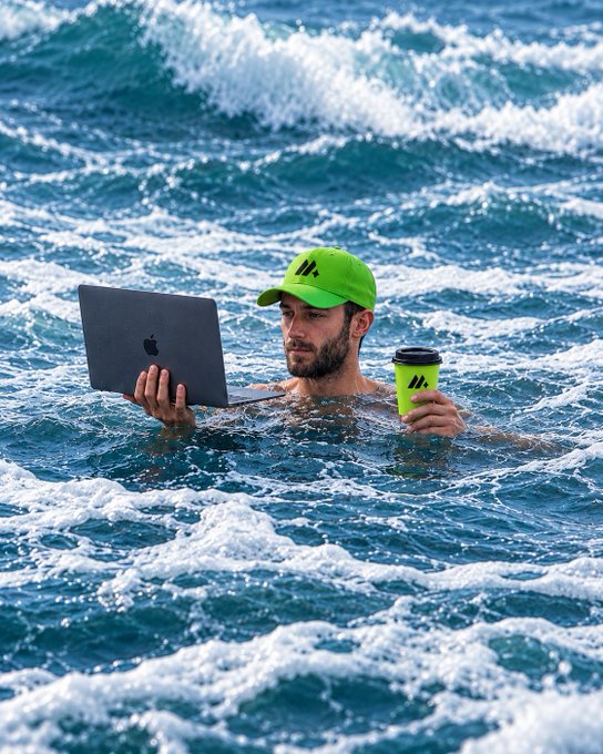 A bearded man in a neon green cap floats amid crashing ocean waves, focused intently on his MacBook while holding a matching green travel mug. The image humorously illustrates the post's point about staying glued to trading charts no matter how chaotic the conditions get—even while battling a stormy sea.