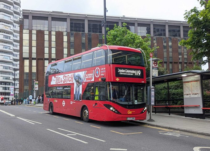 A red London double-decker bus displaying route 119 to 'Trafalgar Square' (via Camden Town) and an 'Lilo & Stitch' advertisement, parked at a bus stop.