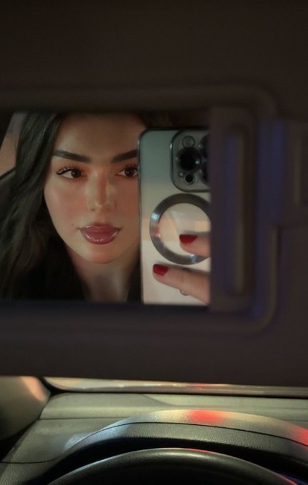 A mirror selfie taken inside a car showing a young woman with dark hair and glossy lips holding an iPhone with a ring grip, with a steering wheel visible below the sun visor mirror.