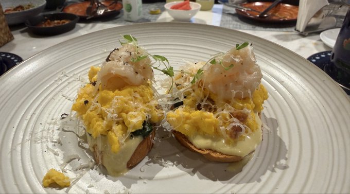 Two servings of eggs Benedict topped with shrimp, grated cheese, and microgreens sit on a white plate.