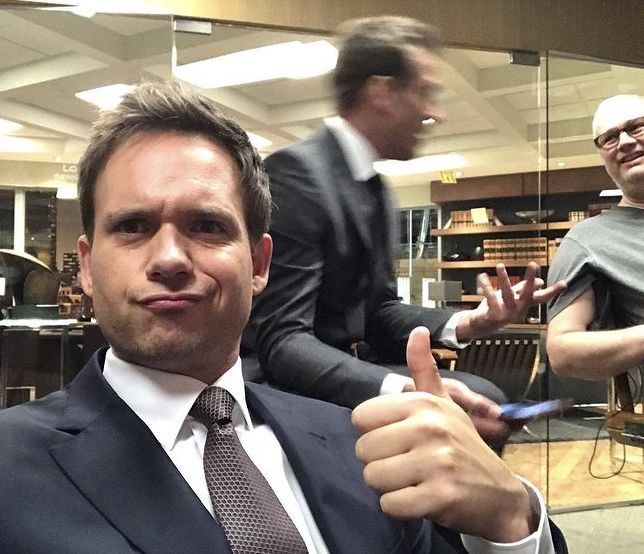 A man in a dark suit and patterned tie gives a thumbs-up to the camera while smirking, with another man in a suit blurred in motion in the background, appearing to be in an office setting.