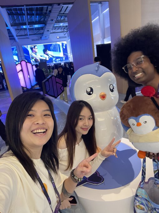 Three women pose for a photo next to a large blue and white penguin mascot at what appears to be a convention or tech event, with one holding a smaller brown plush mascot.
