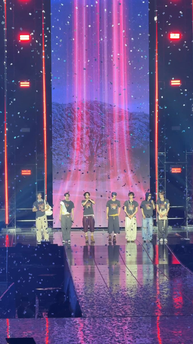 The image features a group of six people standing together, possibly performing or participating in an event. They are positioned in front of a large screen that displays a waterfall scene with red lights falling from the top. The backdrop consists of two large speakers on either side of the screen, and there is also a smaller speaker located at the bottom left corner. The image does not contain any text, but it appears to be related to an event or performance taking place in front of this waterfall display.