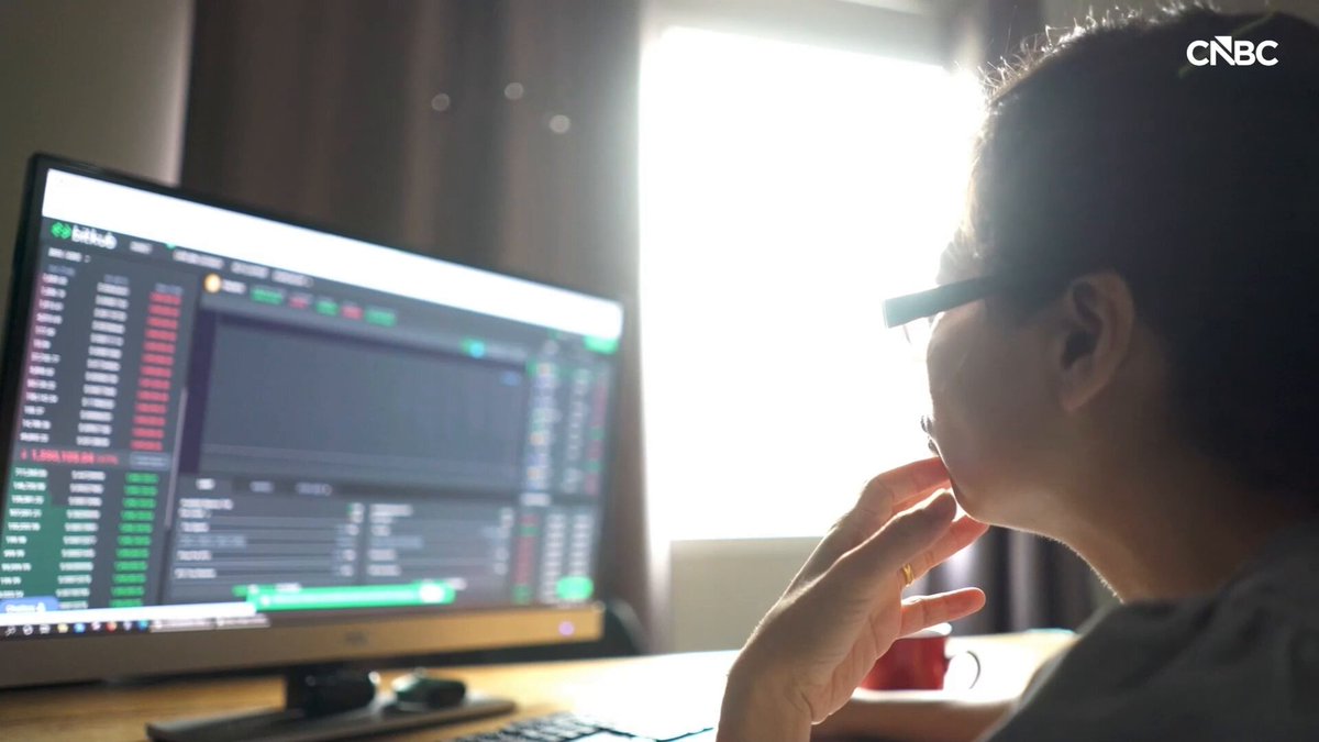 The image shows a person sitting at a desk, looking at a computer screen with a monitor displaying stock market data. The individual appears to be deep in thought or contemplating their next move. The scene suggests that they are engaged in financial analysis or decision-making related to the stock market.
