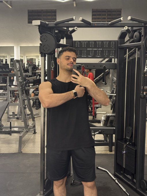 A man in a black tank top and shorts takes a mirror selfie in a gym, standing in front of a cable machine.