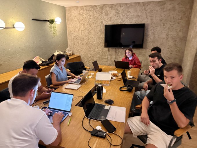 A group of young people sit around a wooden conference table working on laptops, with one man in the foreground eating a sandwich while others type or look at screens.
