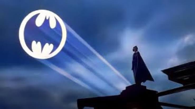 A silhouetted figure in a cape stands on a rooftop against a dark blue sky, gazing up at the illuminated Batman logo projected into the clouds.
