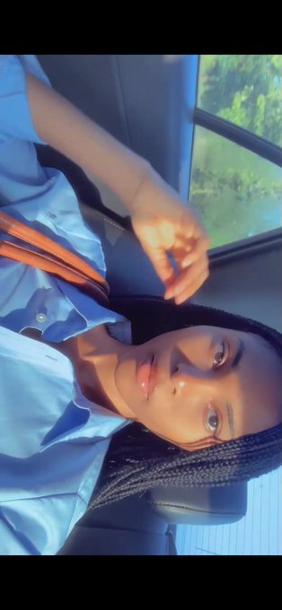 A sideways-oriented selfie of a person with long braided hair wearing a light blue shirt, sitting inside a vehicle with greenery visible through the window.