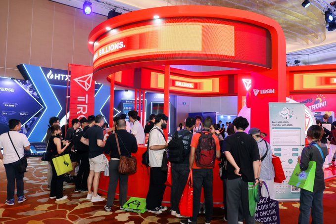 A crowd of people gathers at a red and orange booth labeled 'TRON' and 'MILLIONS,' possibly at a crypto or technology conference with various displays including 'HITX' and 'ZEDON.'