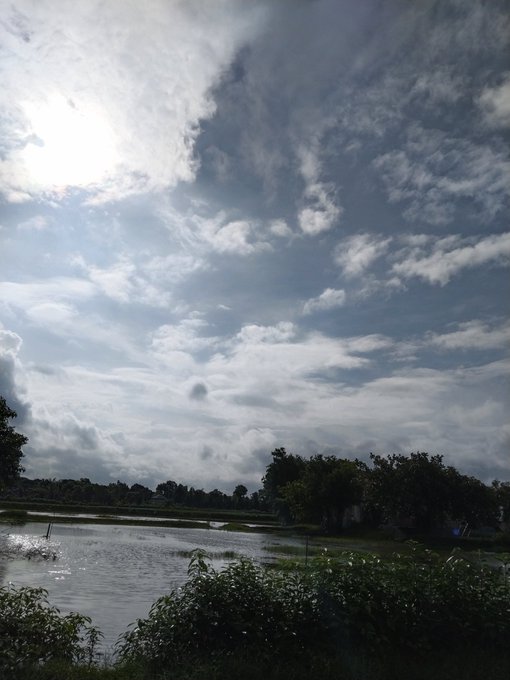 A landscape photograph featuring a dramatic sky filled with scattered clouds over a body of water, likely a pond or flooded field, surrounded by trees and grassy banks.