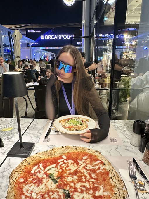 A woman wearing blue reflective sunglasses sits at a restaurant table holding a small pizza, with a larger pizza in the foreground and a busy dining area behind her. In connection with the “solana morning mfers” post, the image appears to capture a casual morning meal; visible text includes a bright “BREAKPOINT” sign and a “solana” logo on the placemats.