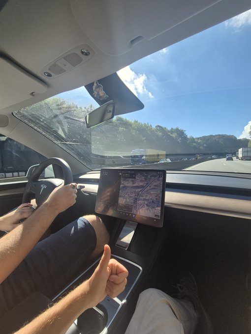 Interior view from the passenger seat of a Tesla on a highway, showing the driver holding the steering wheel while giving a thumbs-up, with the center touchscreen displaying a map/navigation.
