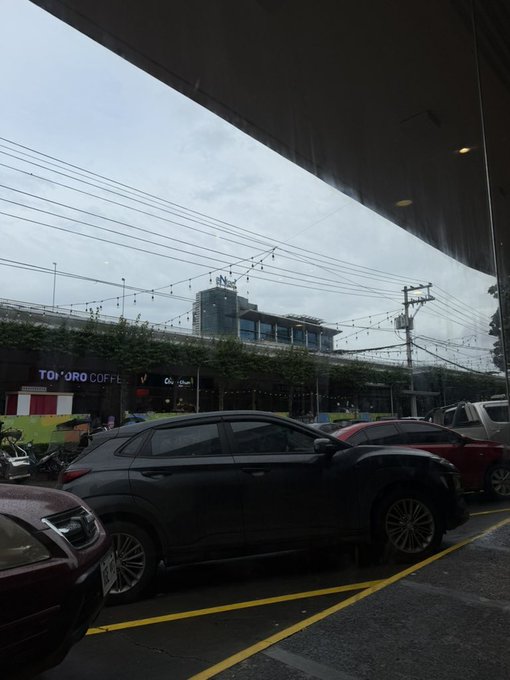 A view from under an awning looking out at a parking lot with several cars, including a dark SUV in the foreground, and a Tororo Coffee shop visible across the street under an overcast sky.