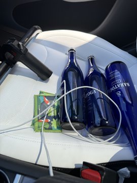 The image shows a car interior with three blue bottles of alcohol, including two wine glasses and one beer bottle, placed on the seat next to a black steering wheel cover. The bottles are arranged in a way that they appear to be connected by a wire or cord, possibly for charging purposes. A red button can also be seen near the center of the image.