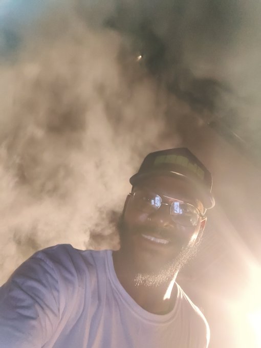 A selfie of a smiling man with a beard wearing glasses, a cap, and a white t-shirt, with dramatic clouds and sky in the background.