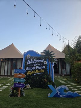 A welcome sign reads "Welcome to Bitget Wallet Night Market" with directional arrows pointing to Bali and a backdrop of white event tents and string lights, capturing the on-site setting of the Bitget Wallet Night Market event mentioned in the post. A decorative blue wave installation sits on the grass in front of the sign, adding to the tropical event atmosphere.