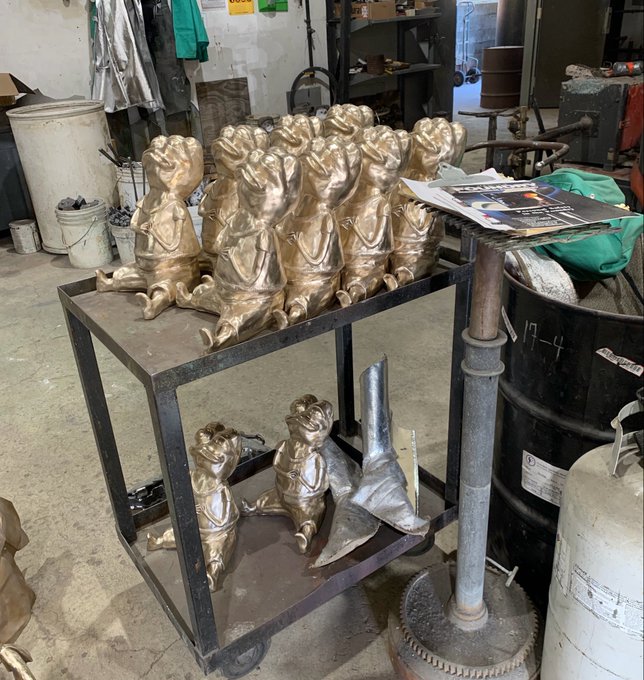 A workshop cart holding several unpainted metal sculptures of frogs or creatures holding flowers, with additional sculptures on a lower shelf amidst various workshop equipment.