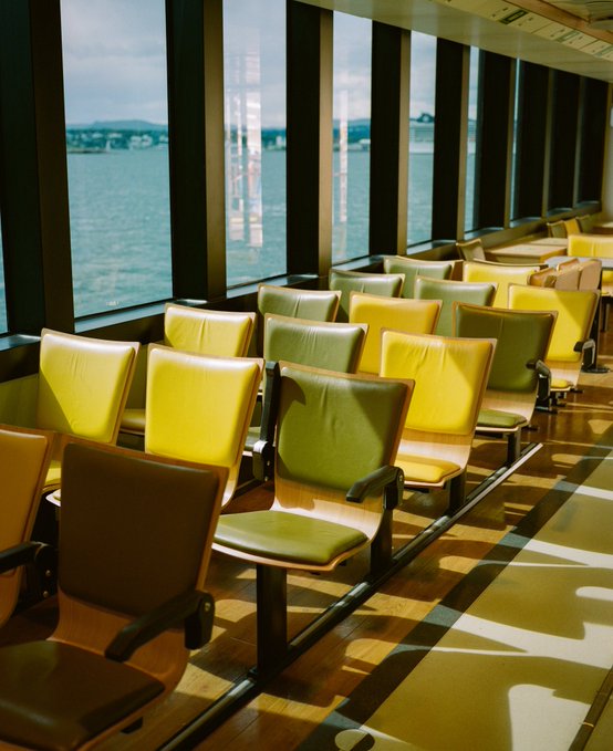 Rows of empty green and yellow chairs line a window overlooking a body of water, with sunlight casting long shadows across the floor.