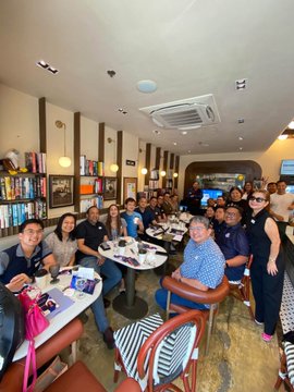A group of people gathered in a restaurant or cafe with wooden chairs and bookshelves, posing for a photo around dining tables.
