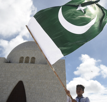 A young boy waves Pakistan's green and white flag featuring the crescent and star emblem against a cloudy sky, with a domed building in the background, symbolizing the intersection of national identity and Islamic tradition relevant to Pakistan's ongoing religious review of cryptocurrency regulations.