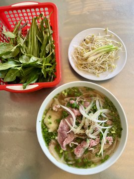 A bowl of Vietnamese pho with sliced beef and onions, accompanied by a red basket of fresh herbs and a plate of bean sprouts on a metal table.