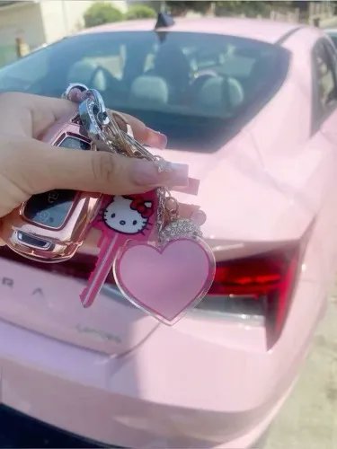 A hand holding a pink Hello Kitty keychain with a heart-shaped charm in front of a pink car, showing the vehicle's "LAVOR" badge.