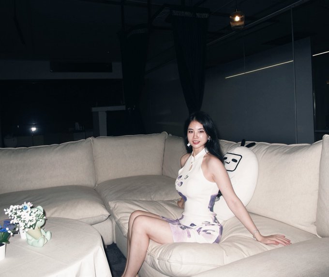 A young woman in a white and purple halter dress poses on a beige sectional sofa in a dimly lit room, with a smiley-face pillow and small flowers beside her. The image appears to contrast with the post, as the youthful photo doesn't match the claim of being 82 years old while introducing herself as a Mixin ambassador and Anders Ventures co-founder seeking connections with market makers and KOLs.