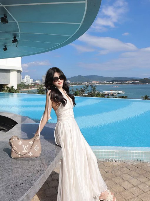 A woman in a flowing white dress and sunglasses poses by a turquoise infinity pool overlooking the Shenzhen coastline, holding a beige Louis Vuitton handbag—reflecting her polished, affluent lifestyle that complements her professional identity as a 25-year-old AI and marketing professional in Shenzhen.