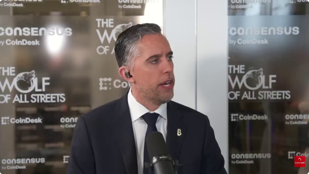 The image shows a man wearing a suit standing behind a microphone at an event or press conference. He appears to be speaking into a microphone, possibly addressing an audience or participating in a panel discussion. The background features a glass wall with white text that reads "THE WOLF OF ALL STREETS" and "CONSENSUS by CoinDesk".