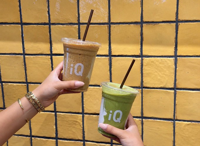 Two hands holding iced coffee and matcha drinks in clear plastic cups with 'iQ' branding, posed in a cheers gesture against a yellow tiled wall.