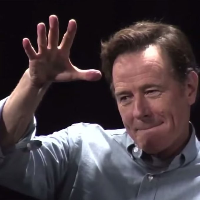 A man with brown hair wearing a light blue shirt smiles while raising his hand with fingers spread against a dark background.