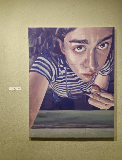 A photograph of an oil or acrylic painting displayed on a pale yellow wall. The artwork is a close-up portrait of a person wearing a black and white striped shirt, leaning forward with an intense gaze and holding something near their mouth.