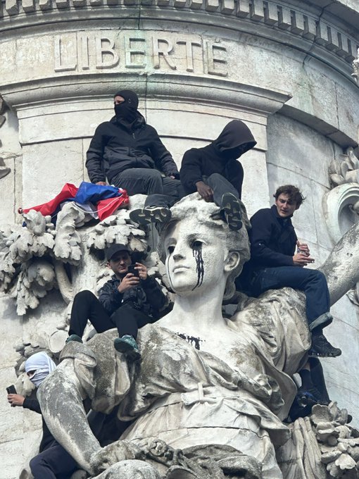 Several masked protesters climb atop an ornate stone monument featuring a sculpted female figure with black paint streaking from her eyes, beneath the word "LIBERTÉ" carved into the architecture—transforming a classical tribute to freedom into a striking scene of contemporary dissent that blurs the line between performance and protest art.