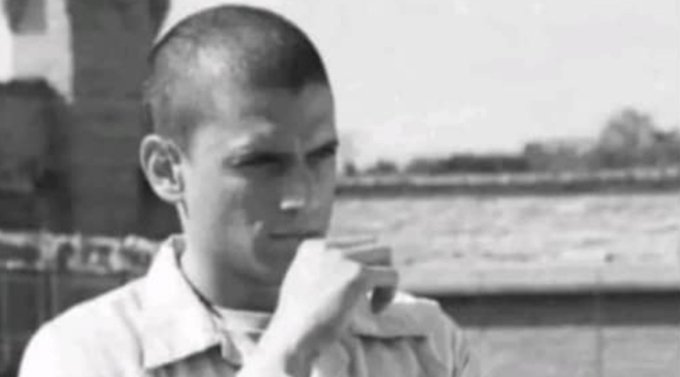 A black-and-white vintage photograph of a young man with a buzz cut eating something outdoors, with water and trees visible in the background.