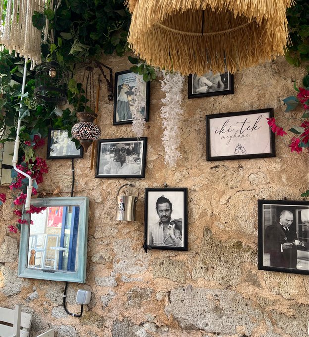 A rustic stone wall display features framed photographs, a 'çhi tek noğlumu' sign, a straw umbrella, hanging plants, and decorative items in a cozy outdoor setting.