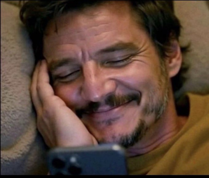 A man with curly hair and a mustache lies down with his eyes closed, smiling contentedly while looking at a smartphone.