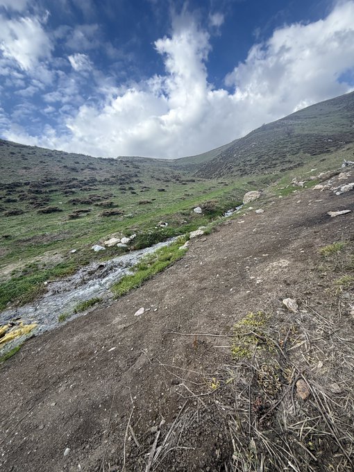 A scenic mountain trail winds through a grassy landscape with a small stream trickling down the slope under a partly cloudy blue sky. The caption "Gm FAM" suggests a cheerful morning greeting to followers, with the peaceful sunrise hike setting embodying a fresh, uplifting start to the day.