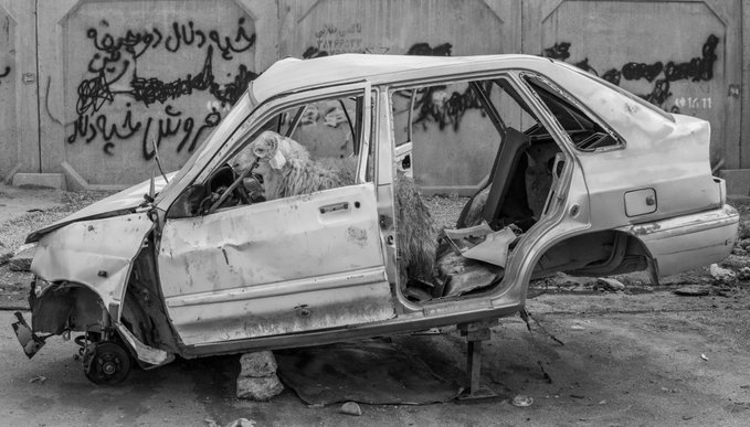 A black and white photograph of a wrecked, stripped car shell propped up on makeshift supports in front of a graffiti-covered wall.