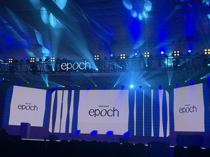 A large event stage is illuminated with blue spotlights, featuring three screens and overhead displays all branded with "sarvam epoch," visually reinforcing the announcement that "Sarvam Epoch is here."