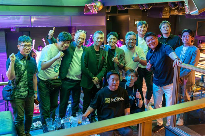 A group of approximately ten smiling men posing together at a bar or lounge venue, with colorful lighting and drinks visible on the counter.