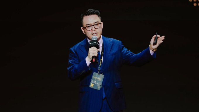 A man in a blue suit and glasses speaks into a microphone while gesturing with a presentation clicker, wearing a "SPEAKER" badge at the Conviction 2026 event, where Tô Trần Hòa, Deputy Head of the Standing Committee of the Department of Crypto Asset Market Management, discussed the five types of assets that could be listed on Vietnam's crypto exchange.
