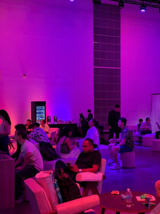 A dimly lit event space bathed in purple and pink lighting, where attendees are seated on white sofas and stools, with a refrigerated beverage station visible in the background.