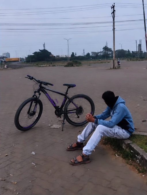 A young man in a blue hoodie and light jeans sits on a curb next to a black mountain bike, looking down at his hands on a paved area near a road.