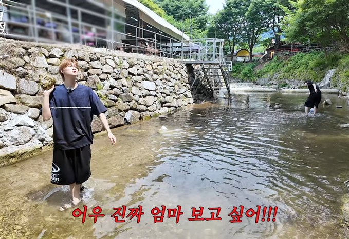 The image shows two people wading in a shallow stream beside a stone wall and walkway, with Korean text overlaid reading "어우 진짜 엄마 보고 싶어!!!" (which translates to "Oh, I really miss my mom!!!"). The #flareucore tag suggests the image is a still from a vlog or video created by the creator flareucore, likely capturing a casual outdoor moment during a stream excursion.