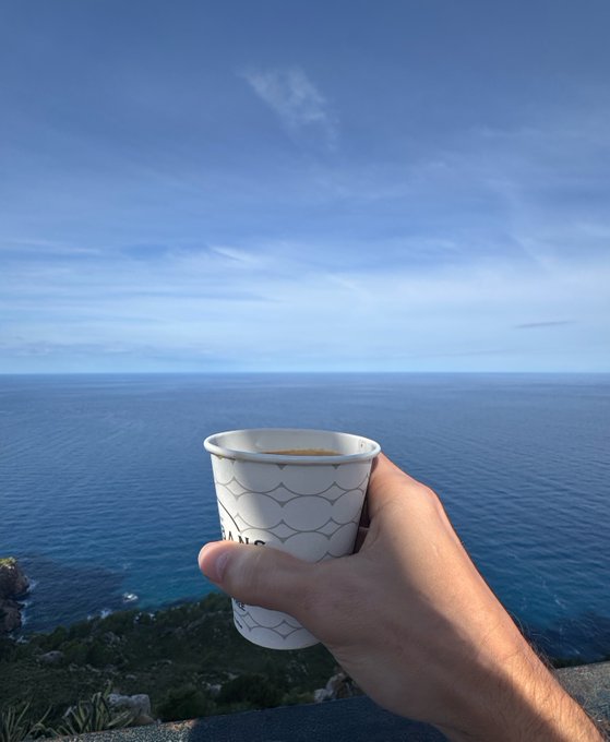 A hand holds a paper cup of coffee in front of a wide blue ocean and pale sky, viewed from a coastal overlook.