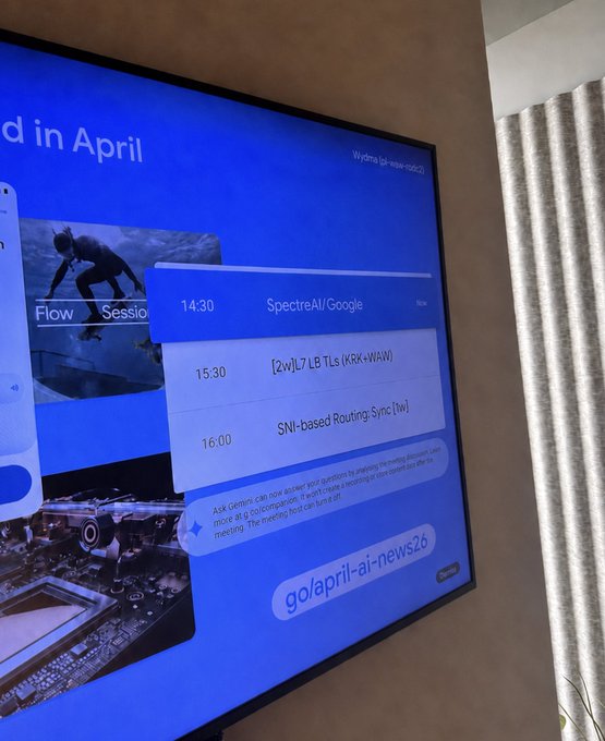 A television screen displays an agenda featuring a 14:30 "SpectreAI/Google" session, alongside entries for L7/L5 TLs (KRK+WAW) at 15:30 and an SN1-based Routing-Sync at 16:00, with a chat box indicating questions can be submitted to go/april-ai-news526—aligning with the $SPECTRE ticker by showcasing the Spectre AI initiative in a technical meeting schedule.