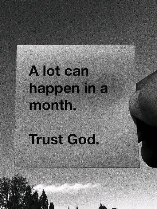A black and white photo of a hand holding a piece of paper with the text 'A lot can happen in a month. Trust God.' against a sky background with trees.