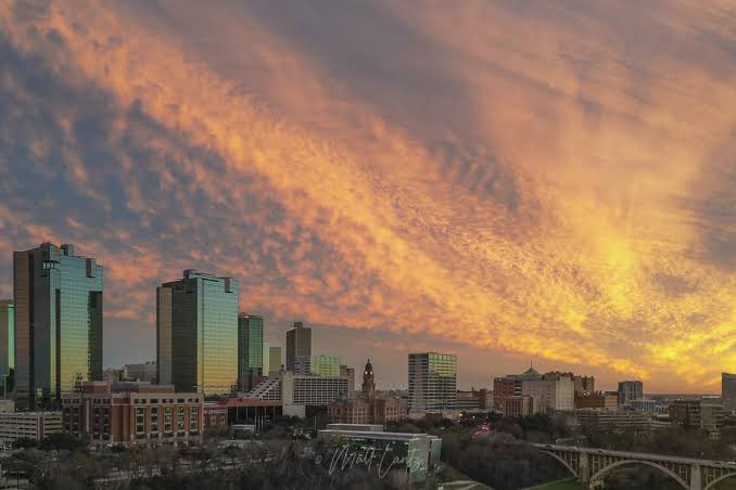 A cityscape at sunset showing modern glass skyscrapers and lower buildings under a dramatic sky filled with rippled orange and pink clouds, with a bridge visible on the right.